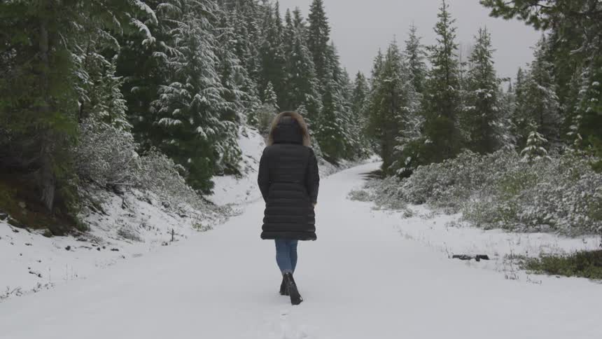 Woman wearing coat walks down on the snowy forest path during daylight