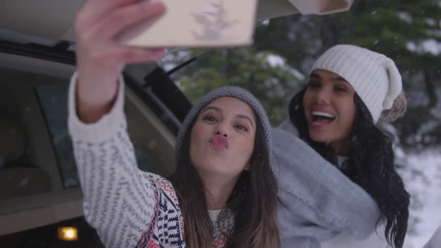 Two young women in winter clothing make kissy lips standing in the falling snow while taking a selfie