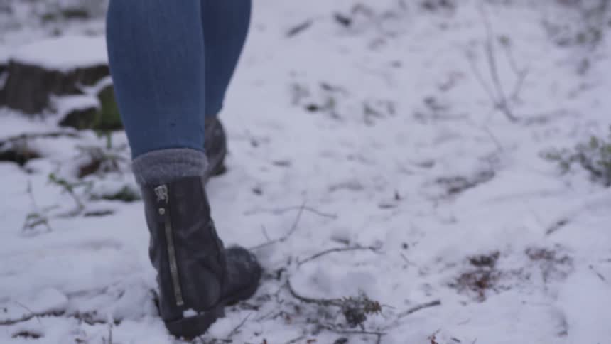 A woman in jeans a black leather boot walks gingerly through the snow on the ground of a wooded area