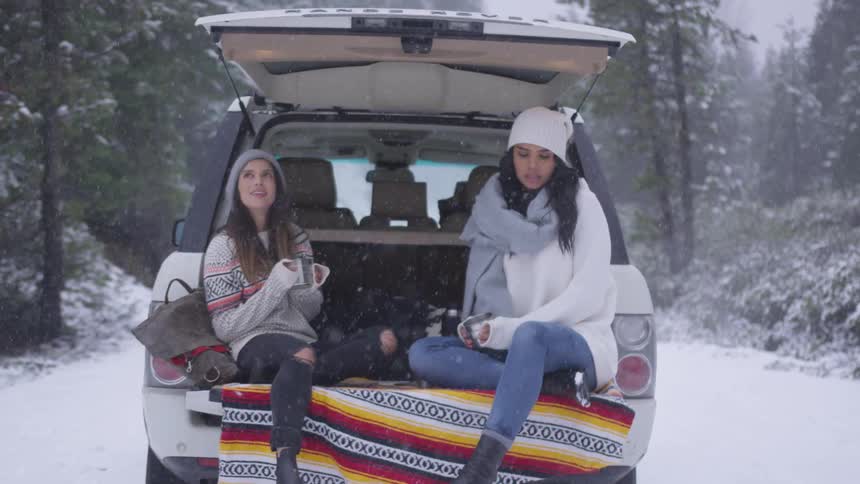 Two young females sit in the back of an open SUV drinking from mugs and watching it snow in a forested area