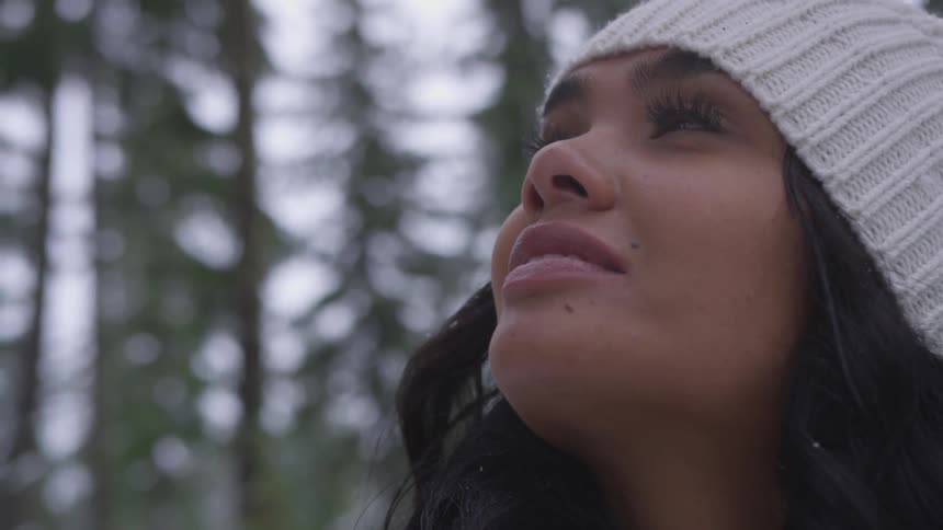 A young woman wearing a white knit hat stands amidst the tall trees of a forest and looks upwards as she smiles
