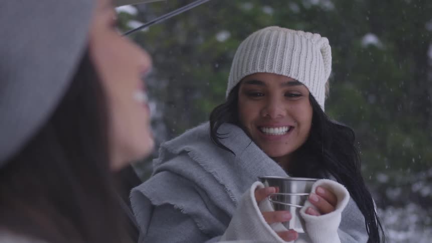 Two women wearing wooly hat smiles and toast with iron mugs in the forest while it snows during daytime