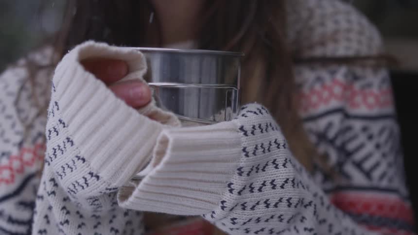 A woman in a sweater is standing outside in the snow and holding a metal cup