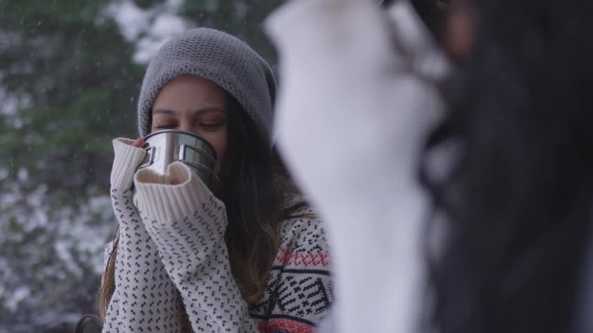 Two women drink from a mug while sitting in the back of a car in the snow