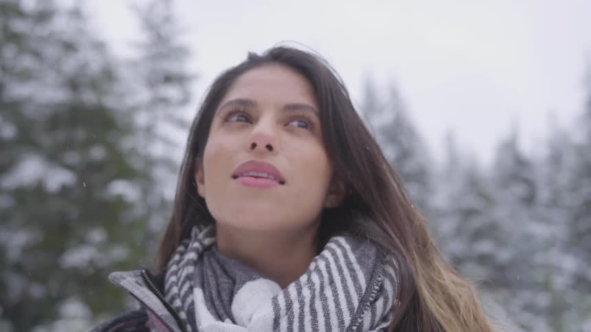 Brunette woman wearing scarf looks around while it snows in the forest during daytime
