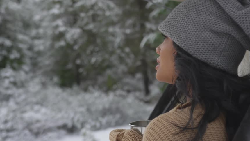 Brunette woman wearing wooly hat sits down on chair and looks ahead while drinking in the snowy forest