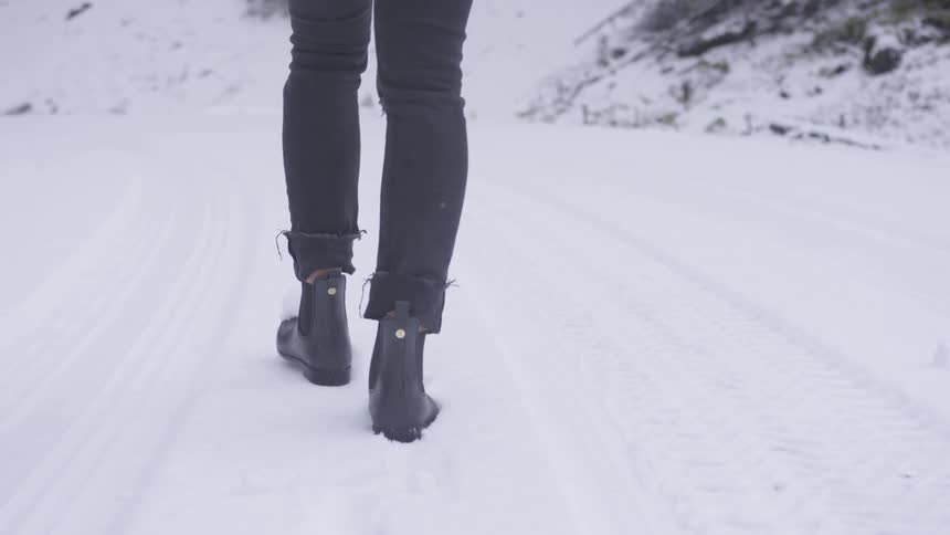 A woman wearing black jeans and rain boots walks through the snow