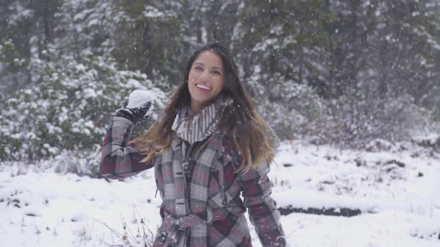 I young adult female in a plaid winter coat throws a snowball at the camera while getting snowed on