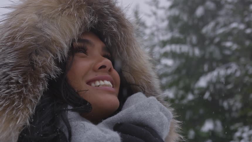 A black woman wearing a winter coat looks up and smiles in a snowy forest