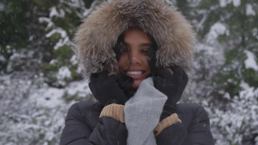 A black woman in winter coat with a fur lined hood smiles while standing in a snowy forest