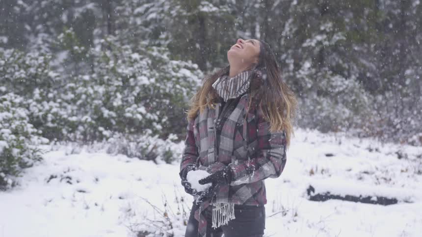 Brunette woman wearing scarf and gloves looks up in the air smiling while snow falls slowly in the forest during daylight