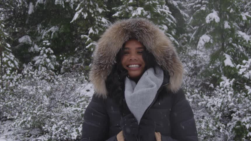 A woman smiles standing outside as it snows by the forest