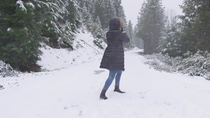 Woman wearing coat walks down on the snowy forest road while it snows in the daytime