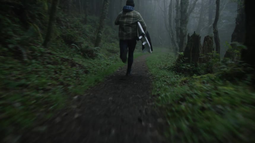 Young man sprints down a foggy forest trail with his surfboard and wetsuit in hand