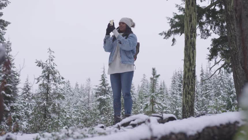 A black woman in a denim coat and a cap uses her smartphone to take a picture in the woods