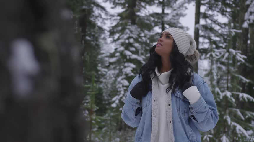 Woman wearing wooly hat and denim looks up in the forest while it snows in the daytime