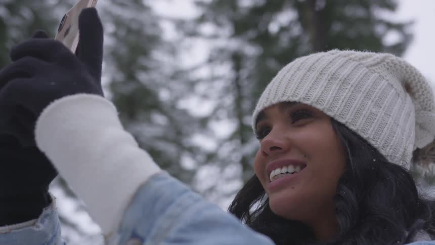A young adult female in a winter hat takes a selfie while small snowflakes land on her with evergreens in the background