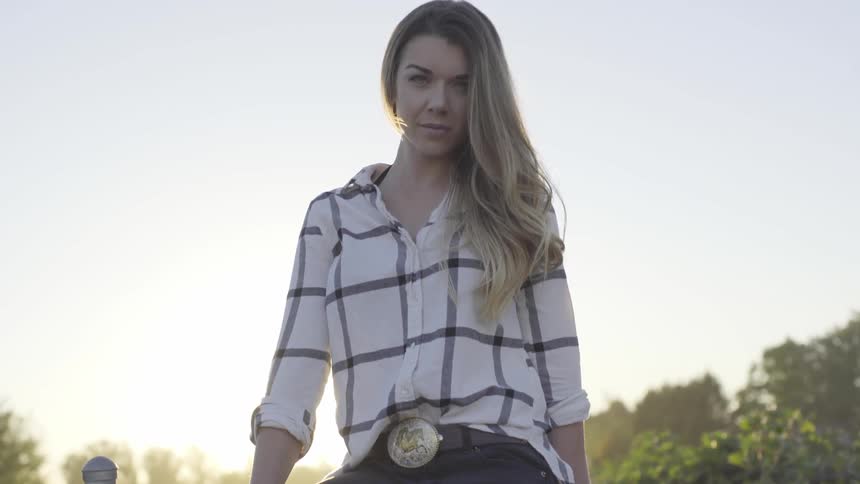 Long blond hair anglo woman wearing a plaid style long sleeve shirt and a cowboy belt buckle turns her head upwards and looks at me