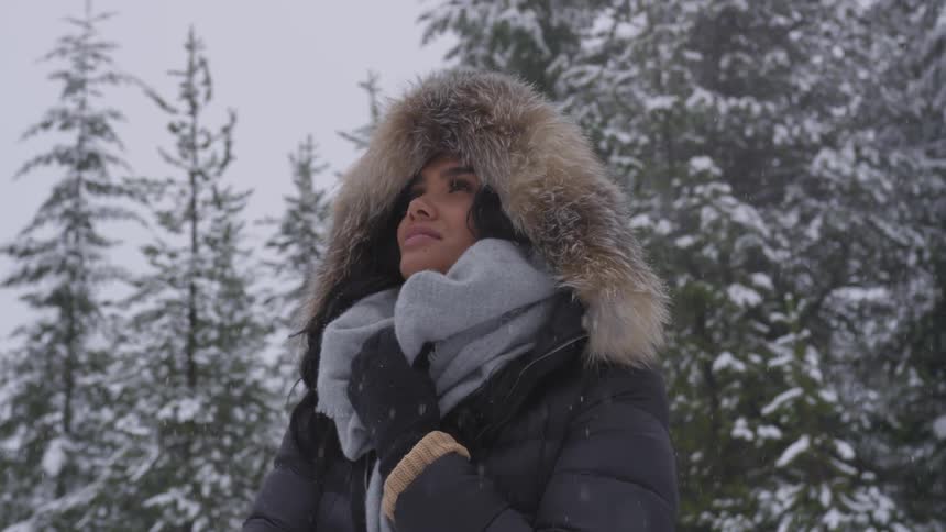 A woman in a heavy jacket watches snowfall in the forest