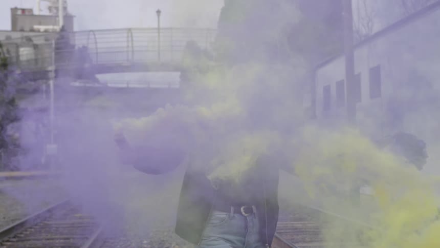 Woman walks down a set of railroad tracks holding yellow and purple smoke bombs