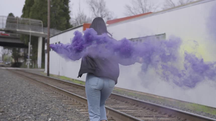 A girl runs in between train track holding colored smoke sticks trailing purple and yellow behind her
