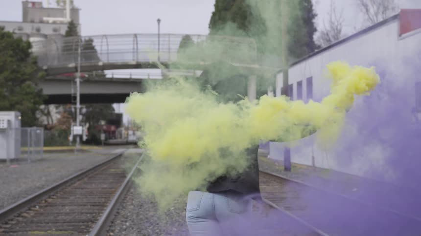 A woman twirls around near train tracks with colored smoke bombs