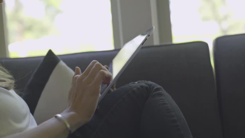 A woman scrolls on a tablet while sitting on a gray couch
