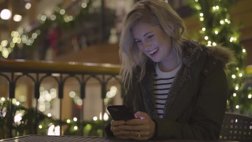 A young white female sits at an outdoor table laughing and smiling while texting on her phone