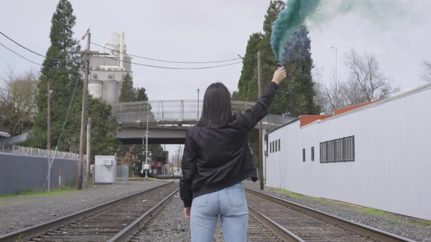A young woman in jeans and a leather jacket holds a blue flare while standing between two railway lines