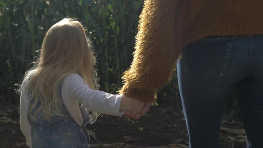 A young girl holds a woman's hand walking through a corn field