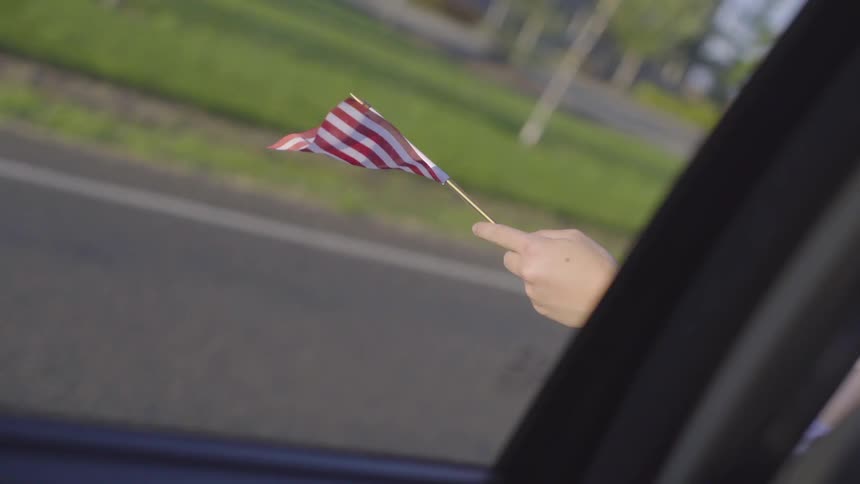 A white person holds a small american flag outside a moving car window