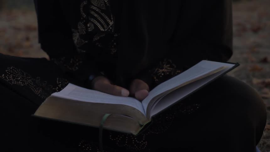 Legs crossed sitting outside on autumn leaves dark clothing hands in lap holding an open book with a ribbon marker