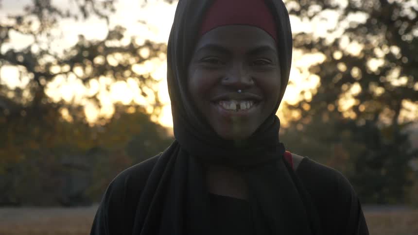 A smiling black woman is wearing a scarf over hear head stands in a clearing in a field surrounded by trees and she shakes her head and smiles very happily