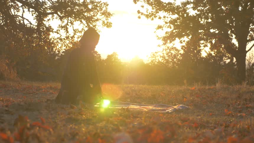 A person spreads a blanket on the ground in a rural field at sunset