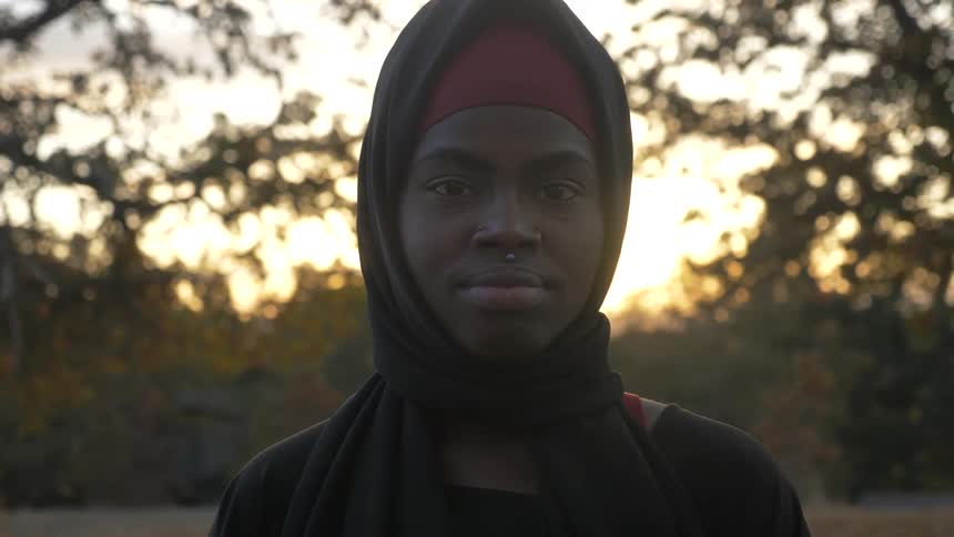 A woman wearing a headscarf stands in front of trees at sunset