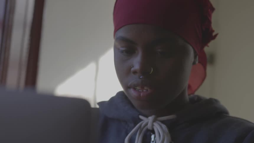 Black girl wearing nose ring and red bonnet uses laptop indoors during daylight