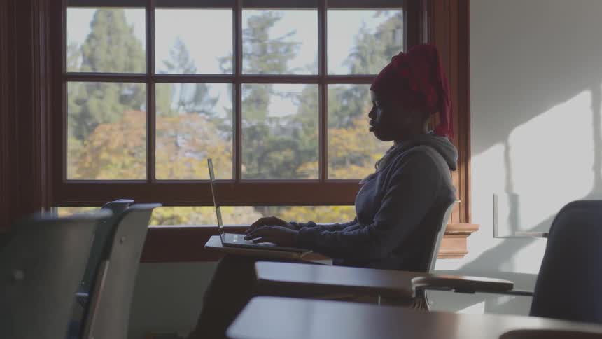 A woman types on a laptop while sitting at a classroom desk next to a window