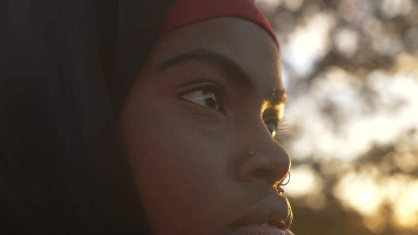 Black girl wearing ring nose and head scarf looks ahead outdoors at sunset
