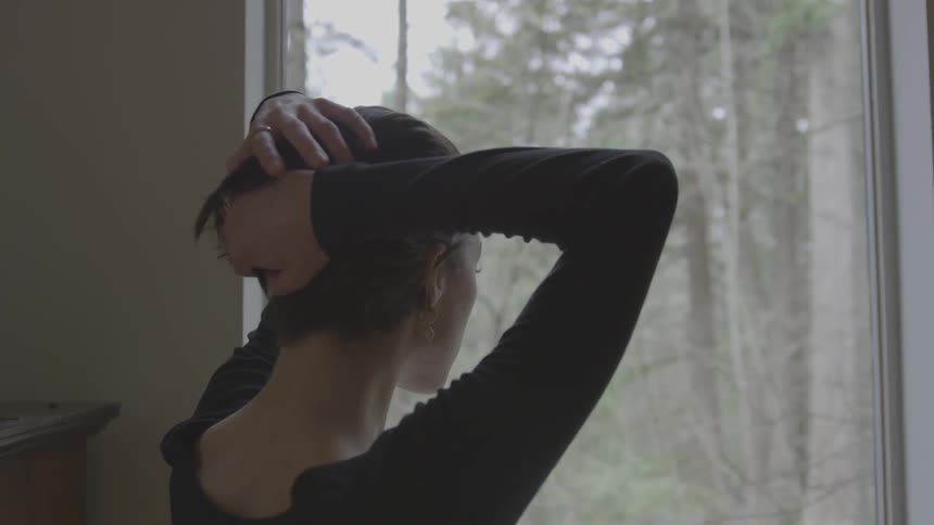 Short haired woman touches her hair while looking out the window on a cloudy day