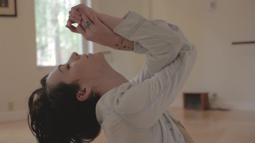 A young white woman with tattoos and wearing a denim shirt over a sweater does yoga