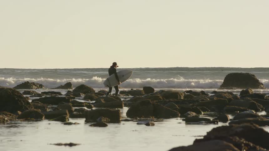 Man holding a surfboard walks on a rocky beach as waves crash in the background