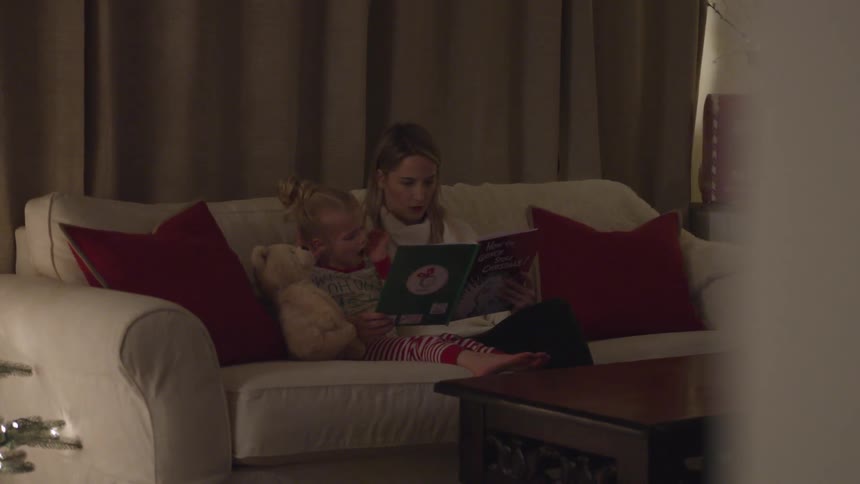 A mother and daughter read a christmas book on a couch by a lit up christmas tree