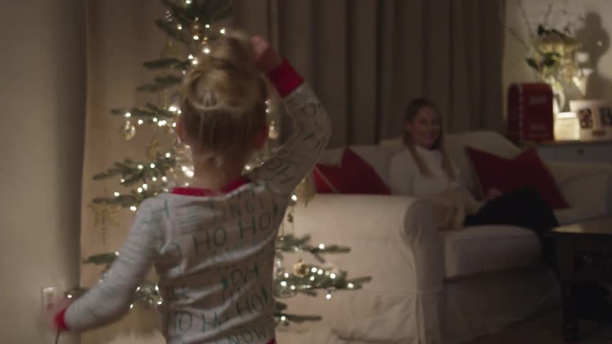 Little girl dances in front of a christmas tree while woman sit on the couch in a dim light room
