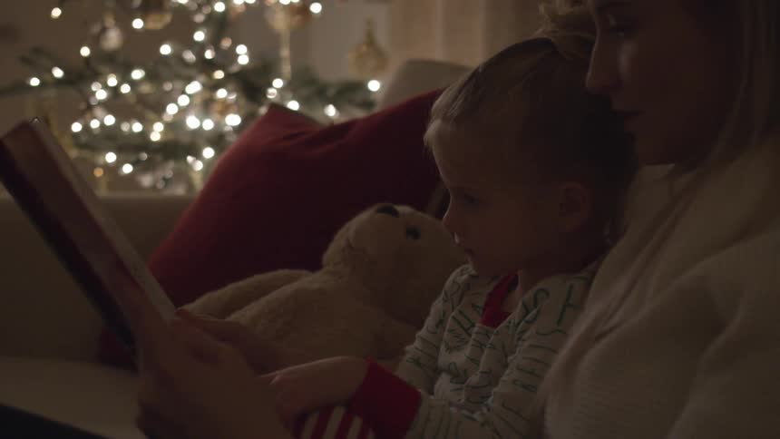 A woman reads a book to a child with a christmas tree twinkling next to them