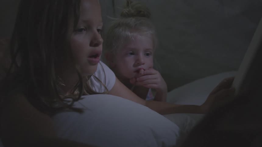 A little girl reads a book to her younger sister while they are both lying on their pillows on a bed