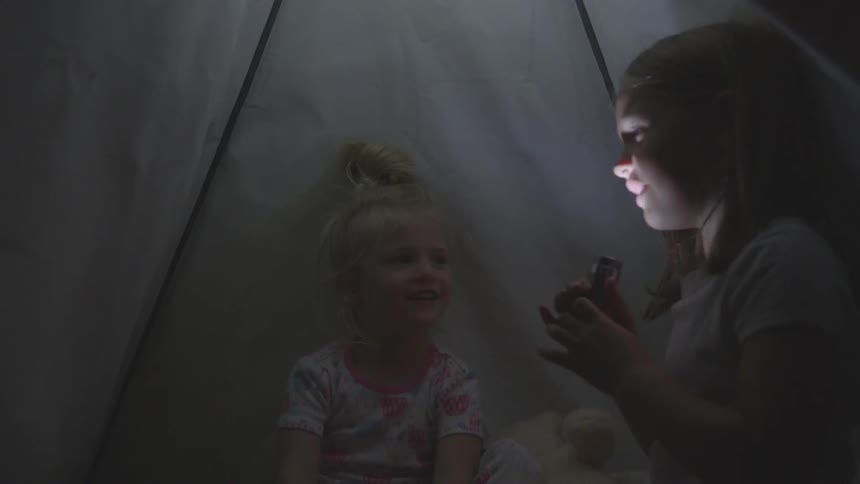 Two young girls sit on a tent while one holds a flashlight