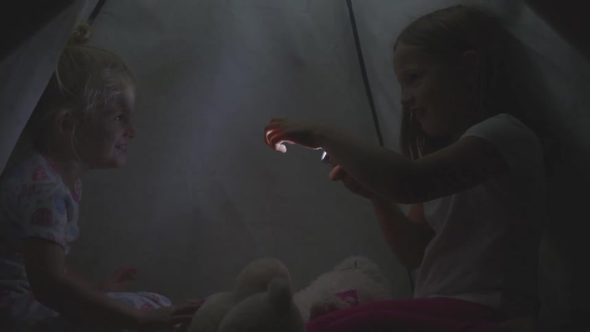 Two young girls laugh and play in a tent with a flashlight