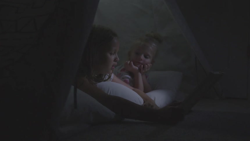 Two little girls lie in a tent looking at a tablet