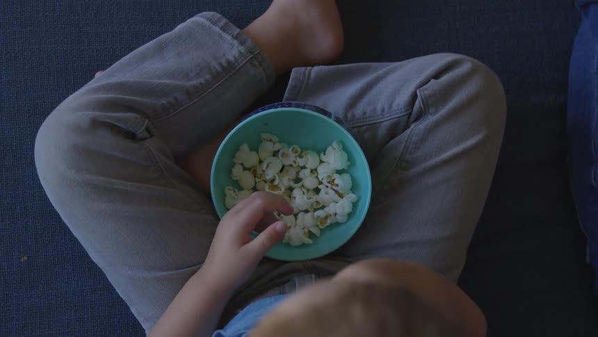 A boy sits and eats popcorn out of a bowl