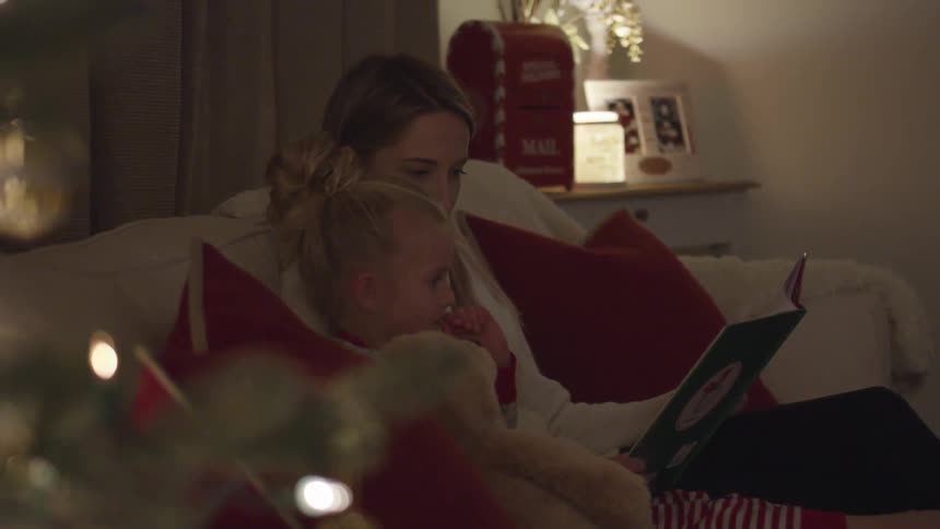 A mother and daughter read a christmas book together on a couch by a christmas tree